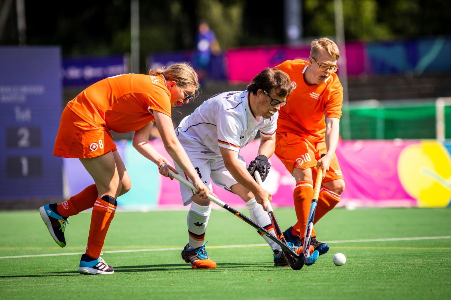 Drei Hockeyspieler, davon zwei im orangenen Trikot, einer im weißen Trikot beim Rasenhockey
