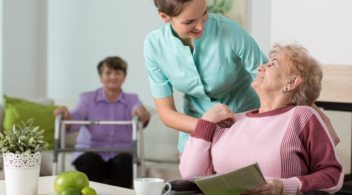 Eine Frau im grünen Kleid beugt sich freundlich zu einer älteren Dame herunter, die am Tisch sitzt. Auf der Sitzbank im Hintergrund eine weitere Frau mit Rollator