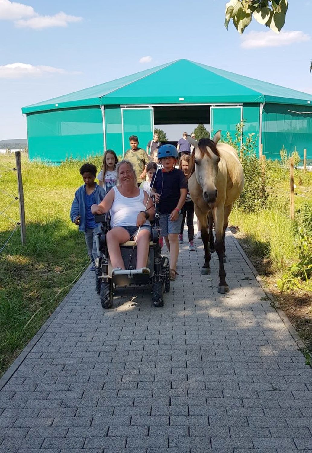 Frau im Elektrorollstuhl auf einem Plattenweg von einem grünen Zelt im Hintergrund. Dabei wird sie von Kindern mit Schutzhelmen begleitet