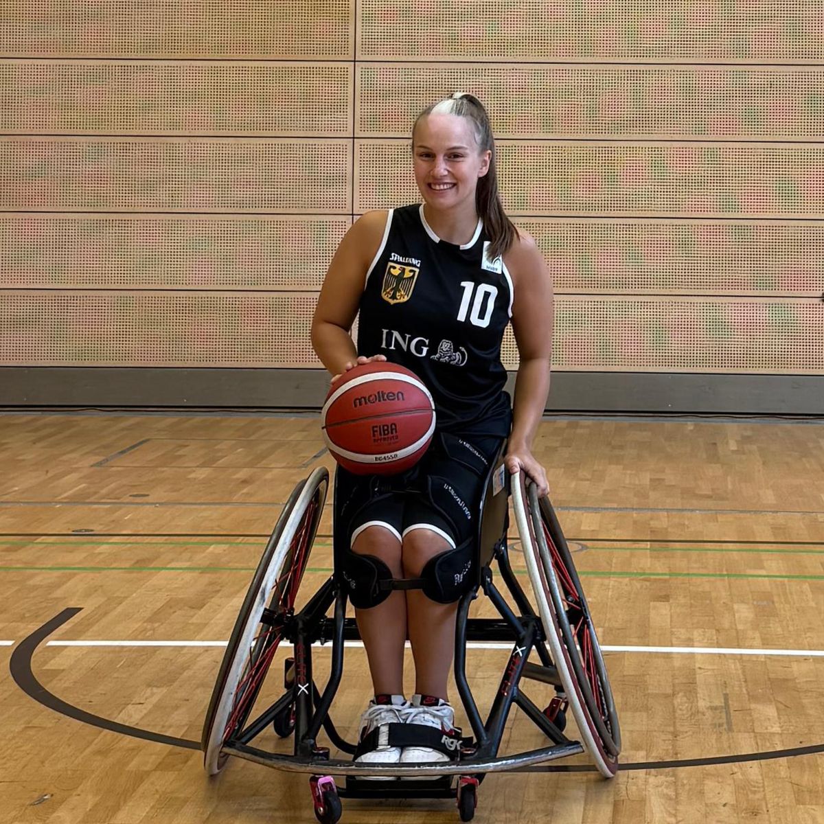 Junge Frau mit langen dunklen Haaren in einem Sportrollstuhl mit einem Volleyball auf den Knien