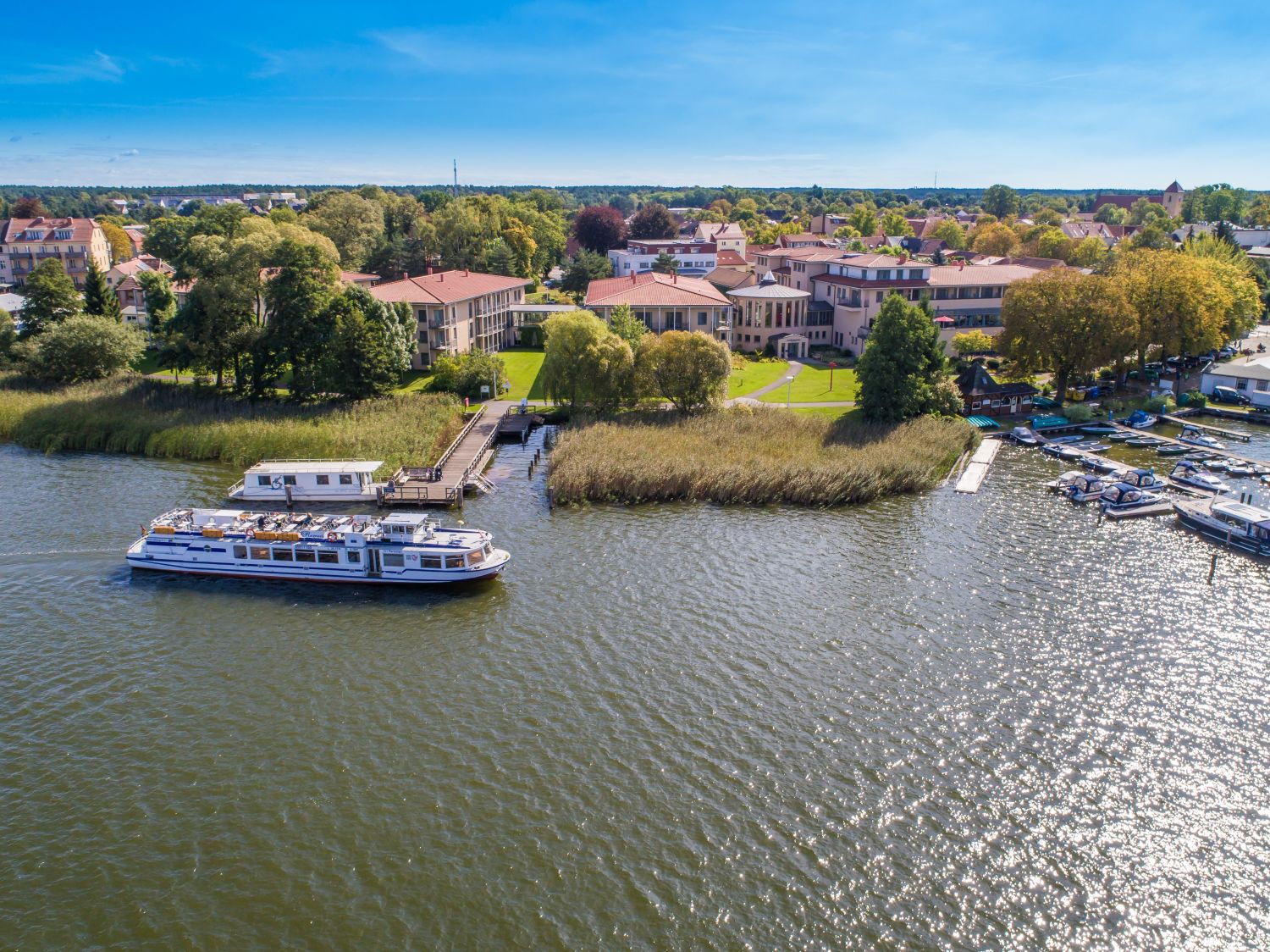 Unterer Teil des Bildes zeigt einen See auf dem sich ein Passagierschiff befindet. Der obere Teil des Bildes zeigt einen Blick auf die Stadt Rheinsberg mit dem Rundbau des Seehotels.