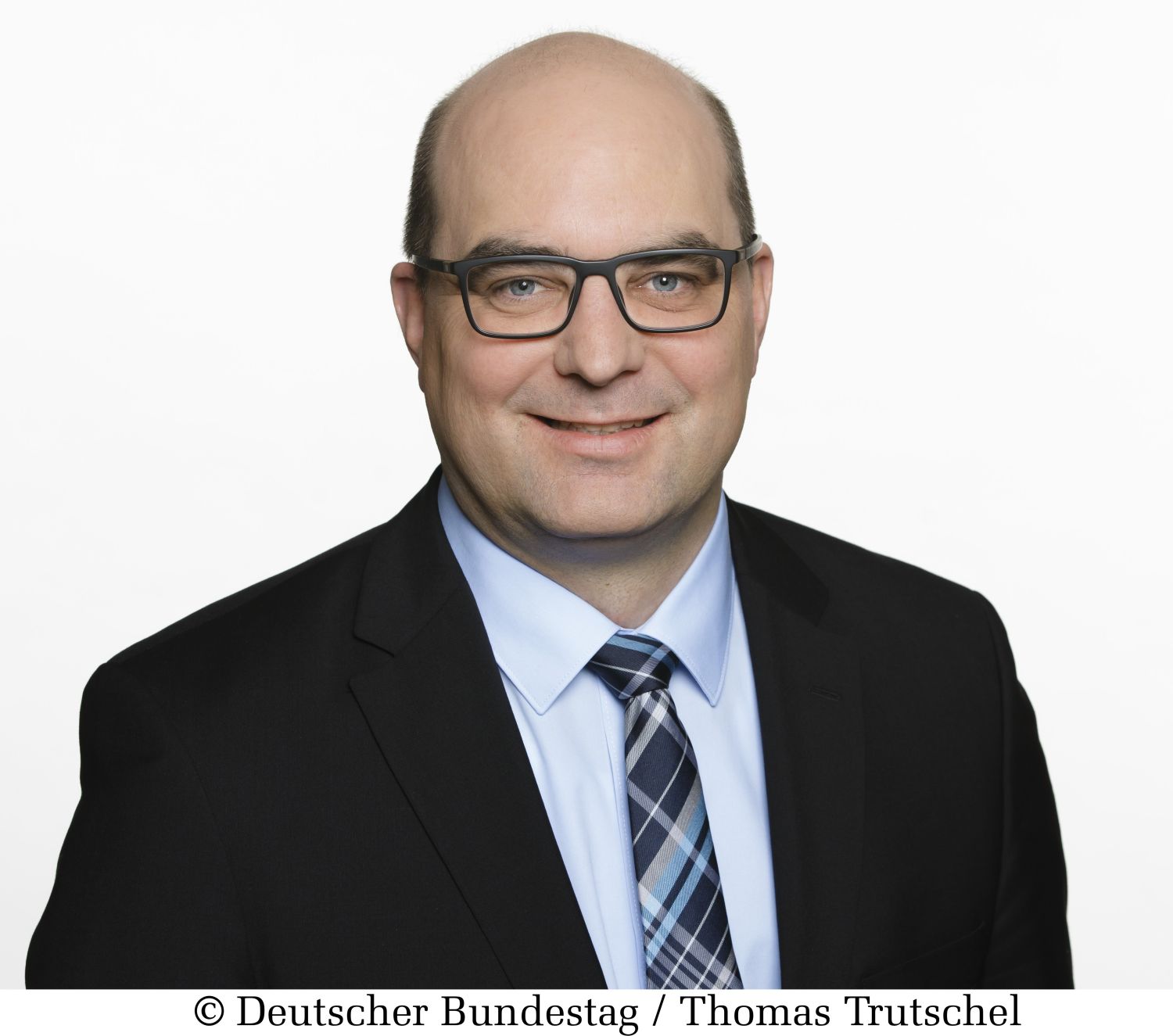 Mann mit dunkler Anzugjacke, hellem Hemd und dunklen karriertem Schlips. Er trägt eine Brille mit dunklem Rand und Haarwuchs an den Seiten des Kopfes