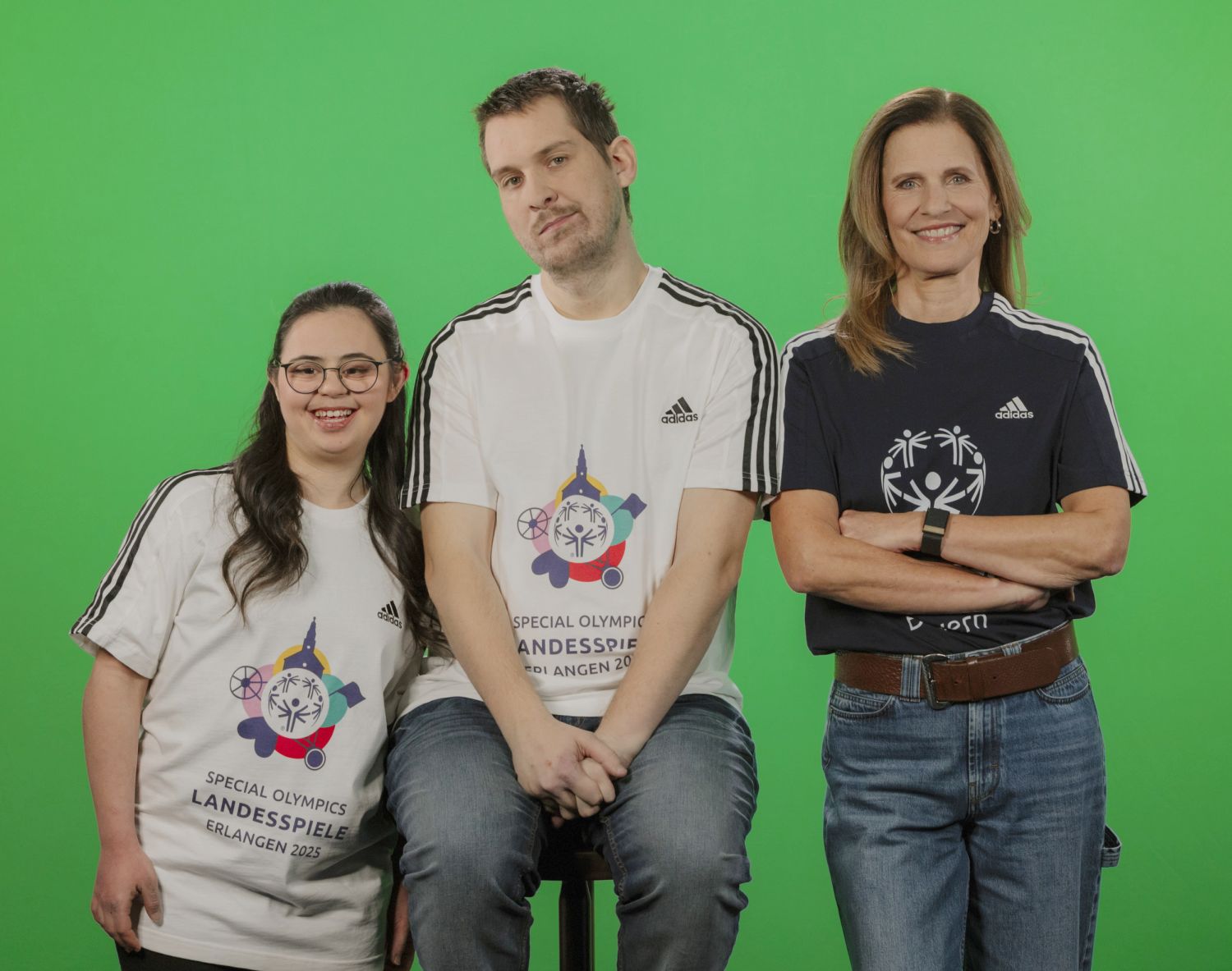 Zwei junge Frauen und dazwischen ein junger Mann, der auf einem Hocker sitzt. Zwei tragen ein weißes, die Frau rechts ein dunkles T-Shirt