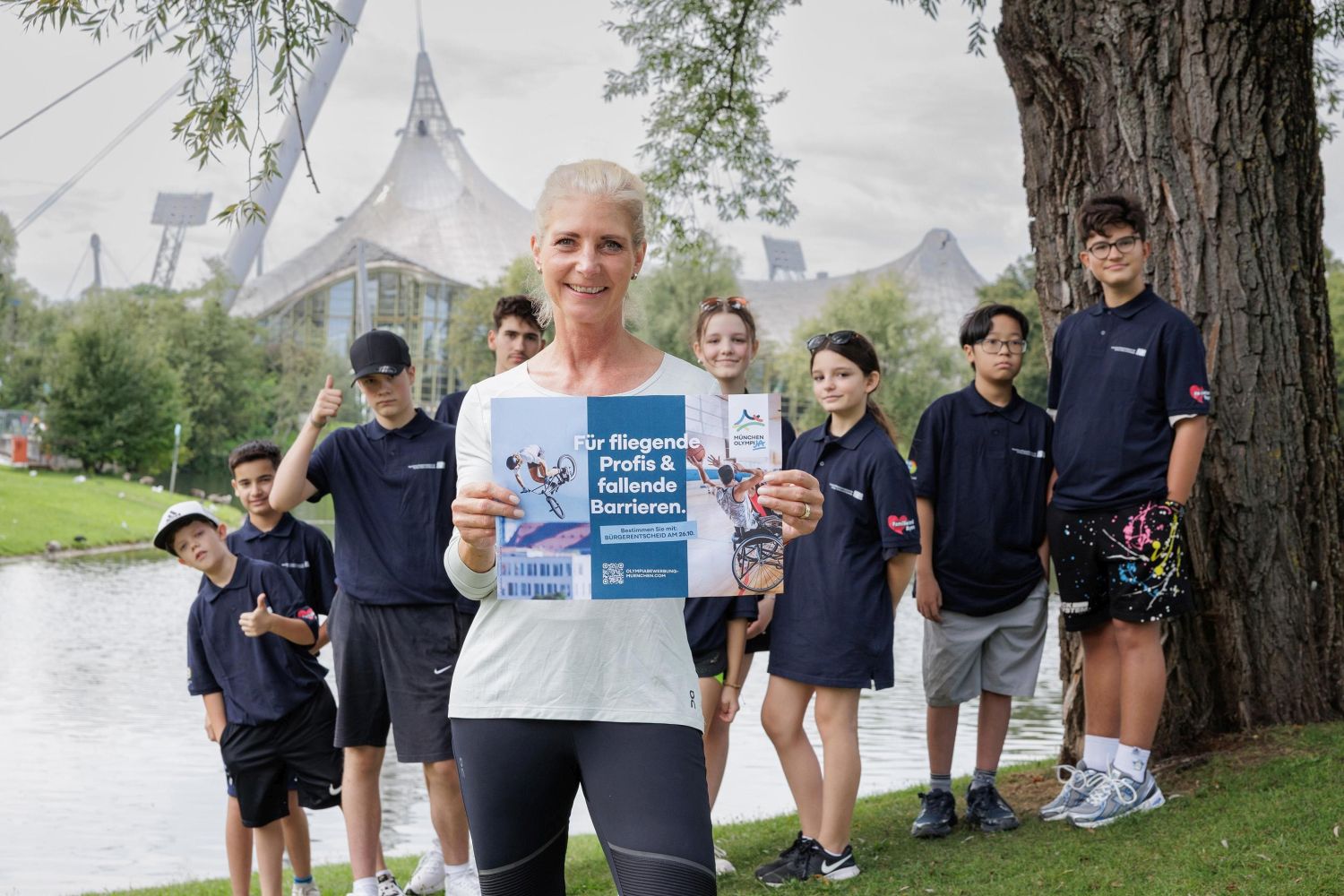 Eine Frau mit weißem T-Shirt im Vordergrund, dahinter acht jugendliche mit blauen Hemden. Im Hintergrund ein Teil des Olympiastadions