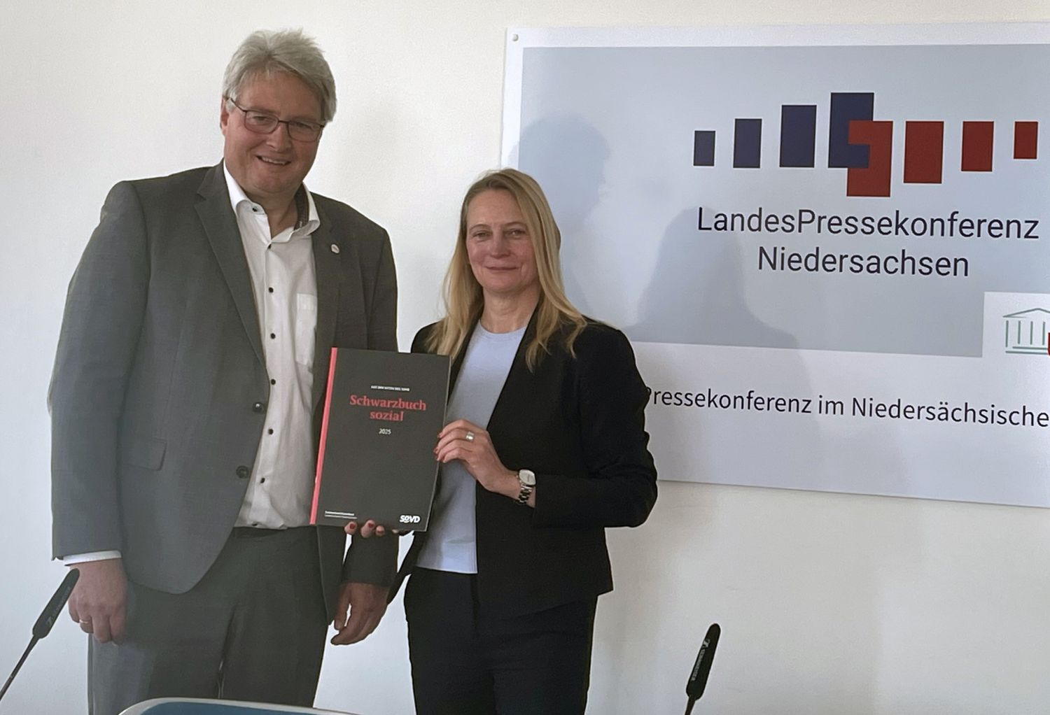 Ein Mann und eine Frau von etwas kleinerer Statur stehen an einer Wand mit der Aufschrift 