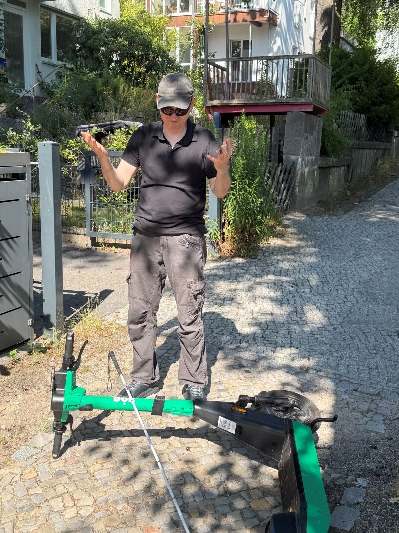 Ralf Rohmann hält empört mit beiden Händen seinen Kopf. Vor ihm liegt ein umgefallener Roller, auf dem ein Blindenstock liegt.