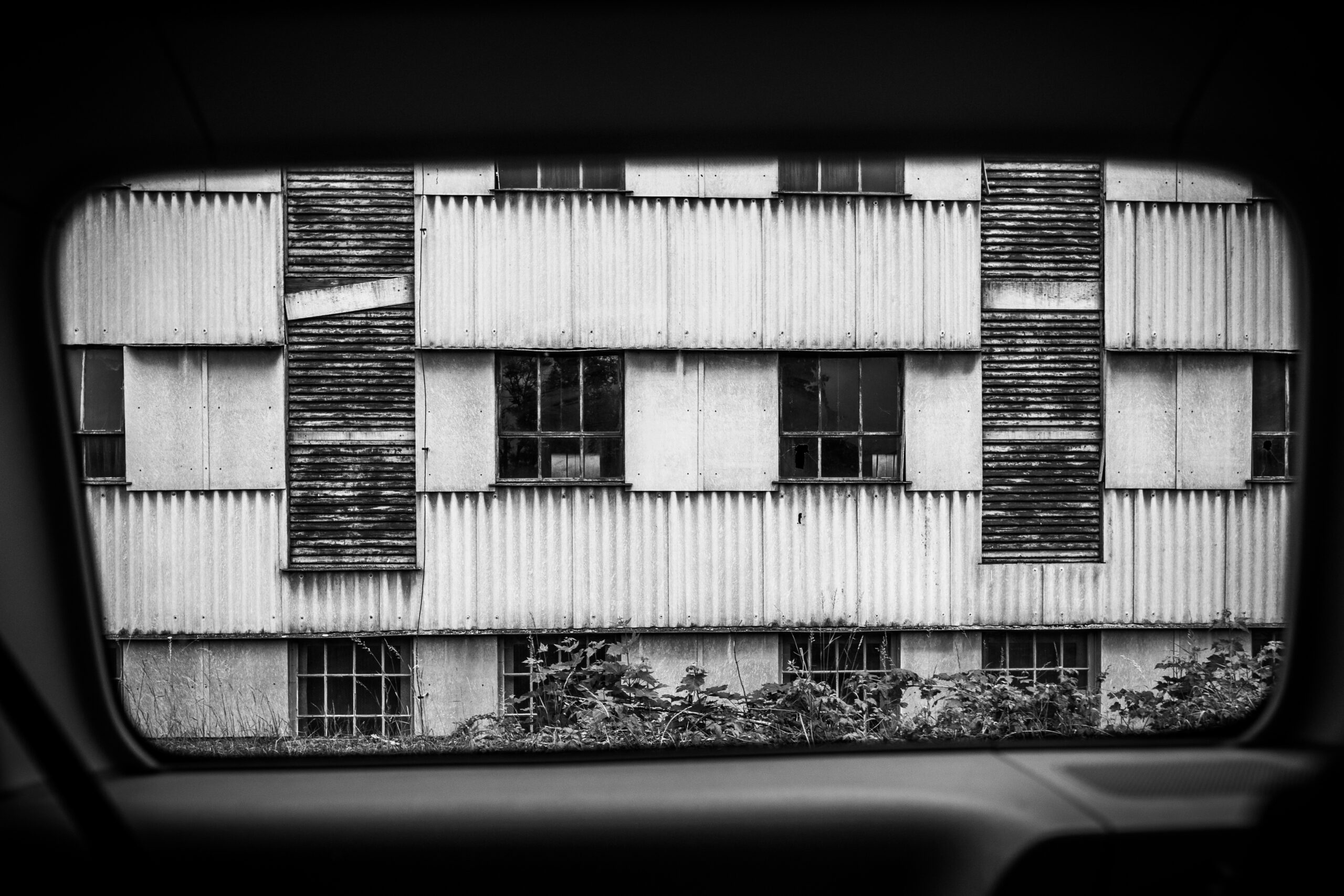 eine verwitterte Fassade, gesehen durch ein Autofenster. Starr, blockhaft, funktional.