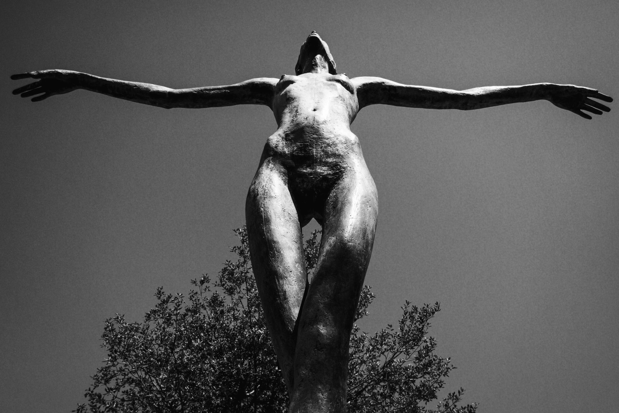 Bronzeskulptur eines aufgerichteten, nach oben gestreckten menschlichen Körpers mit weit ausgebreiteten Armen. Die Aufnahme zeigt die Figur aus der Untersicht, vor hellem Himmel und einem angedeuteten Baum im Hintergrund.