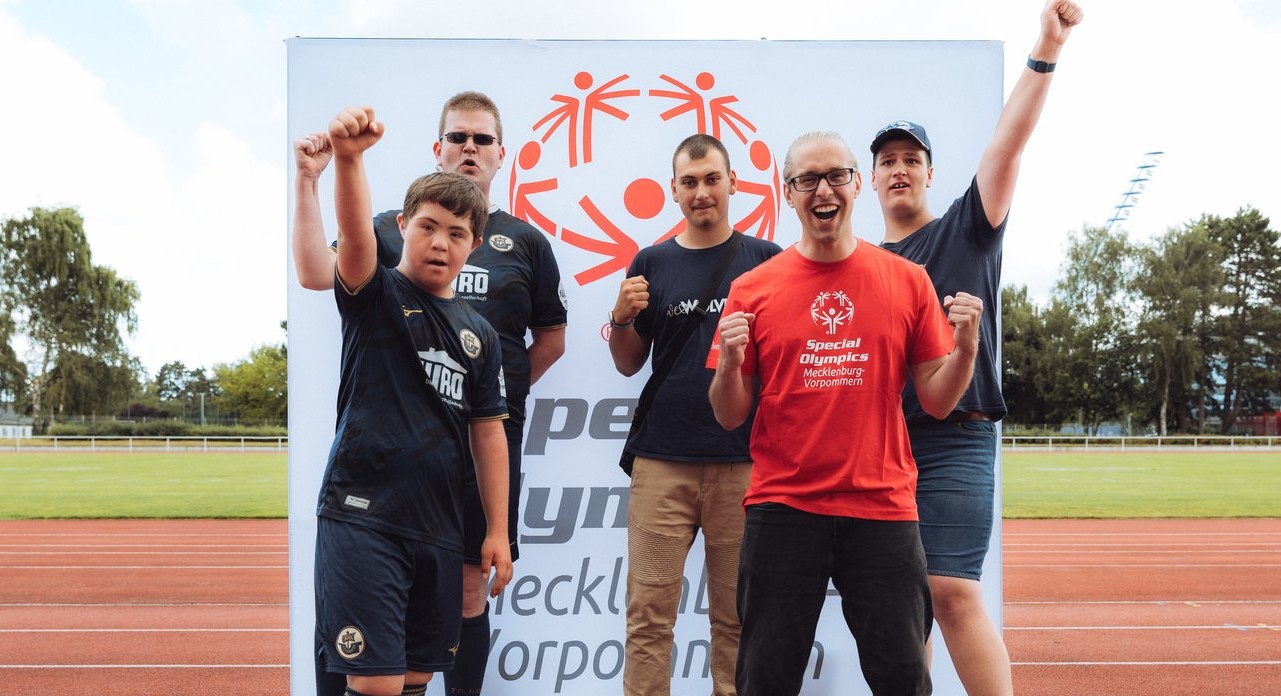 Fünf Sportler mit unterschiedlichem Sportdress vor einer großen Tafel mit dem Signet der Special Olympics