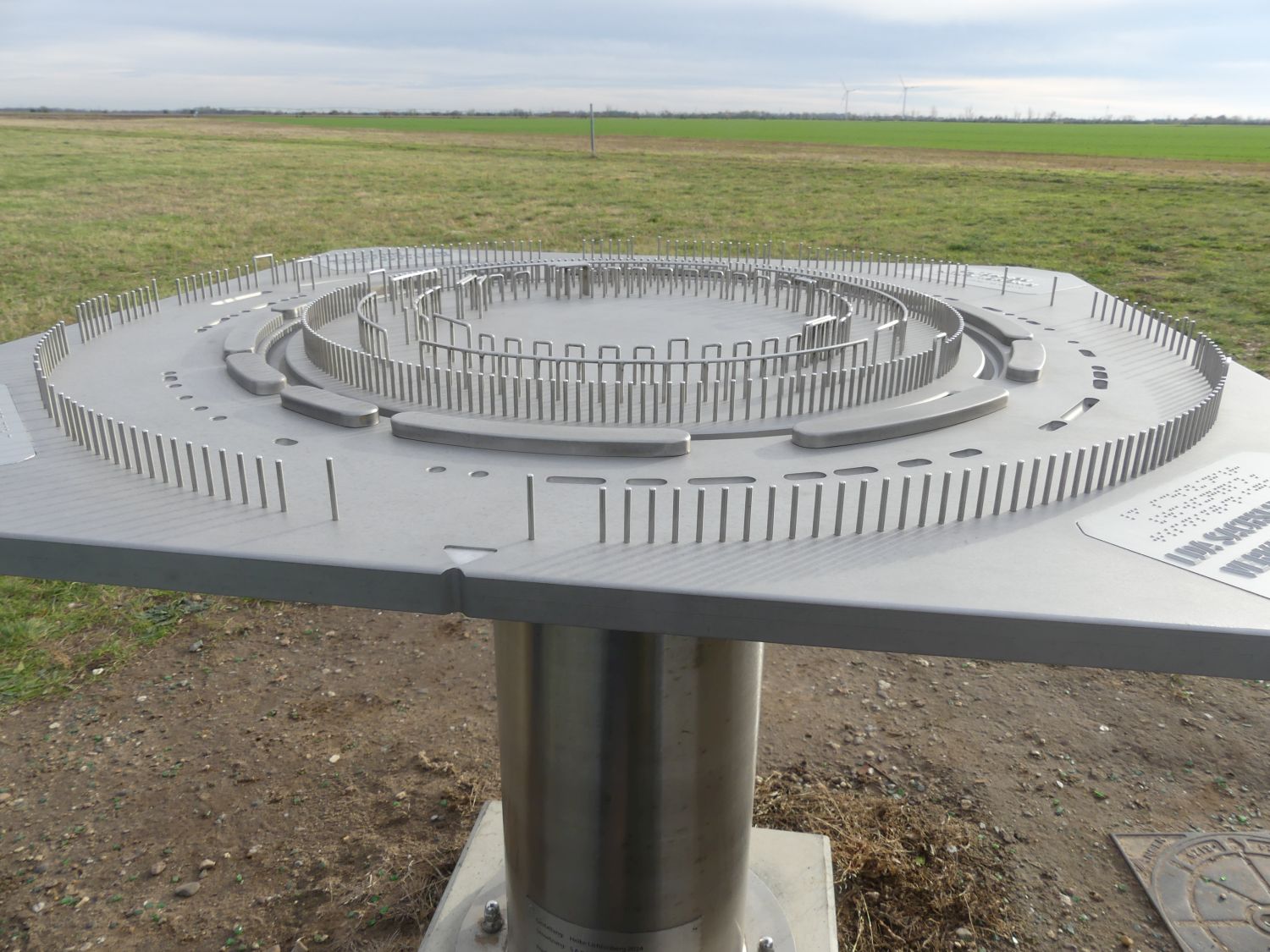 Quadratische Metallplatte die auf Tischhöhe in Ringanlage Pömmelte mit ihren Pfosten und Wegen zeigt