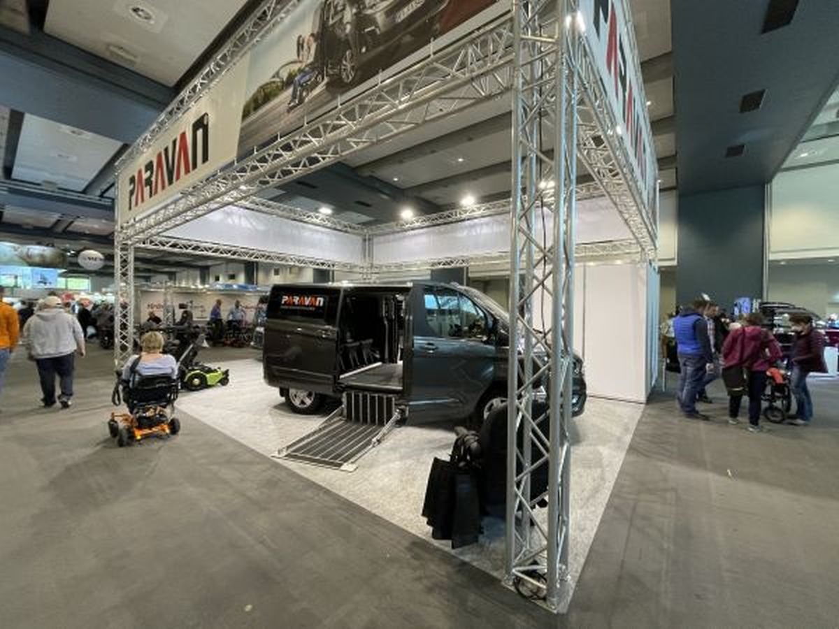 Messestand mit einem dunklen großen Auto mit geöffneten Türen. Am Messestand sind Besucherinnen und Besucher mit und ohne Rollstuhl zu sehen