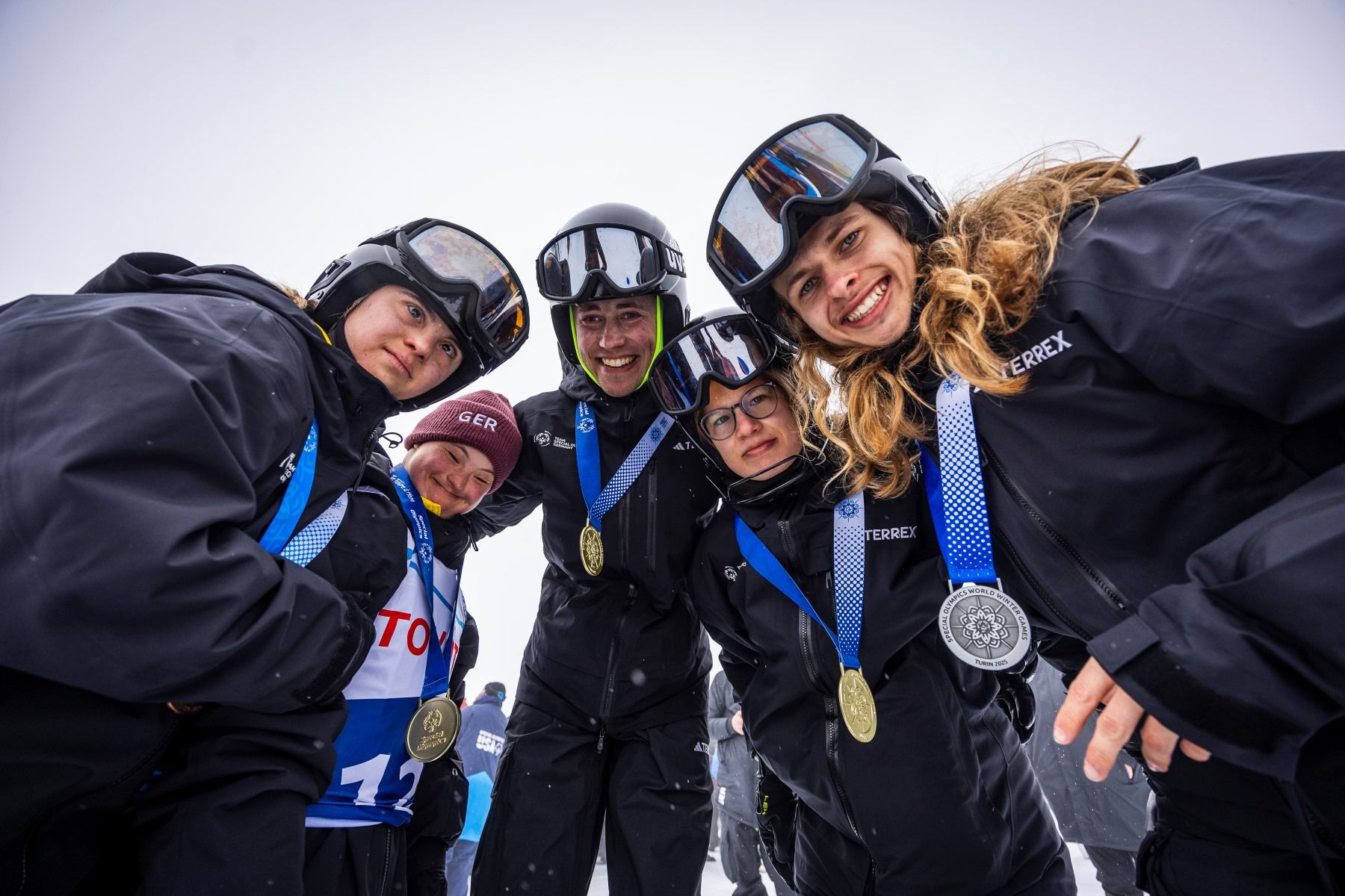 Sportlerinnen und Sportler in dunklen Anzügen beugen sich über die Kamera und zeigen ihre Medaillen