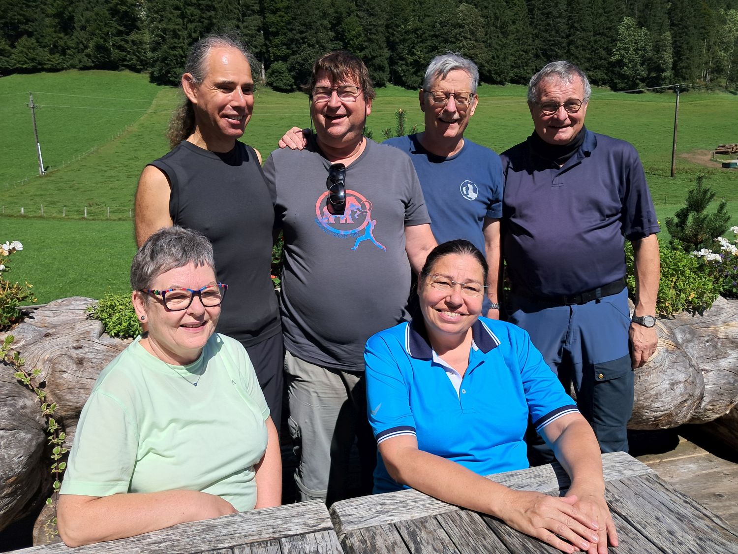 zwei Frauen sitzen an einem einfachen Tisch an einer Baknk. Dahinter stehen vier Männer. Sie alle tragen einfache Wanderkleidung. i, Hintergrund ist Wald zu sehen